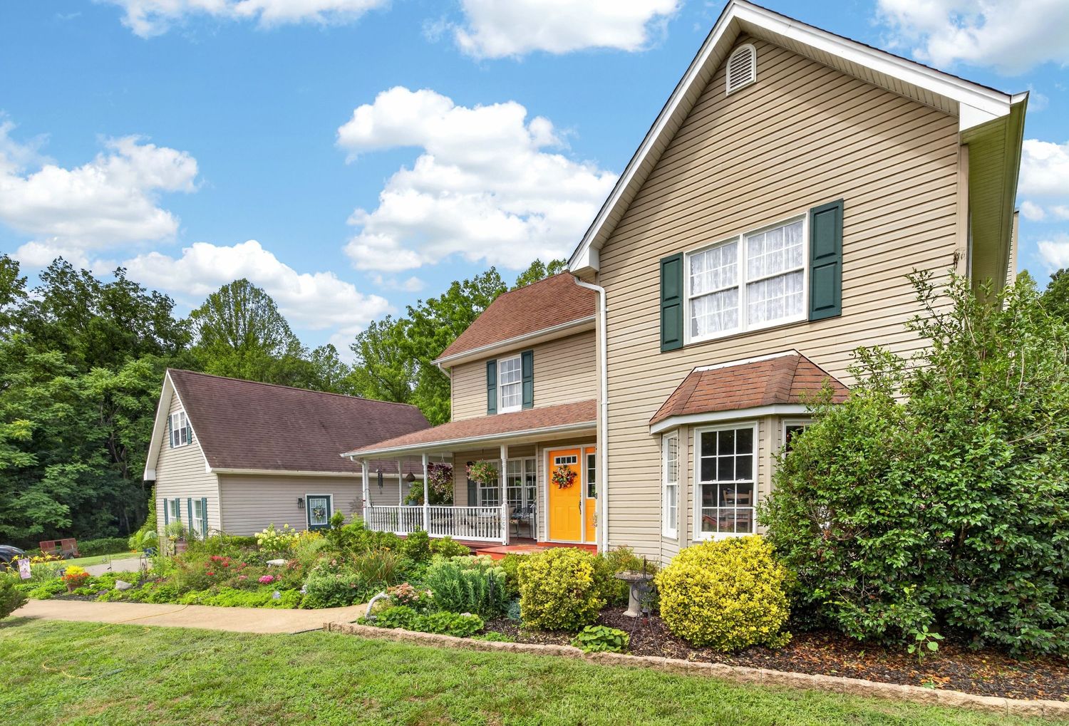 Two-story colonial home with tan siding, green shutters, covered porch, and manicured landscaping surrounded by lush trees.