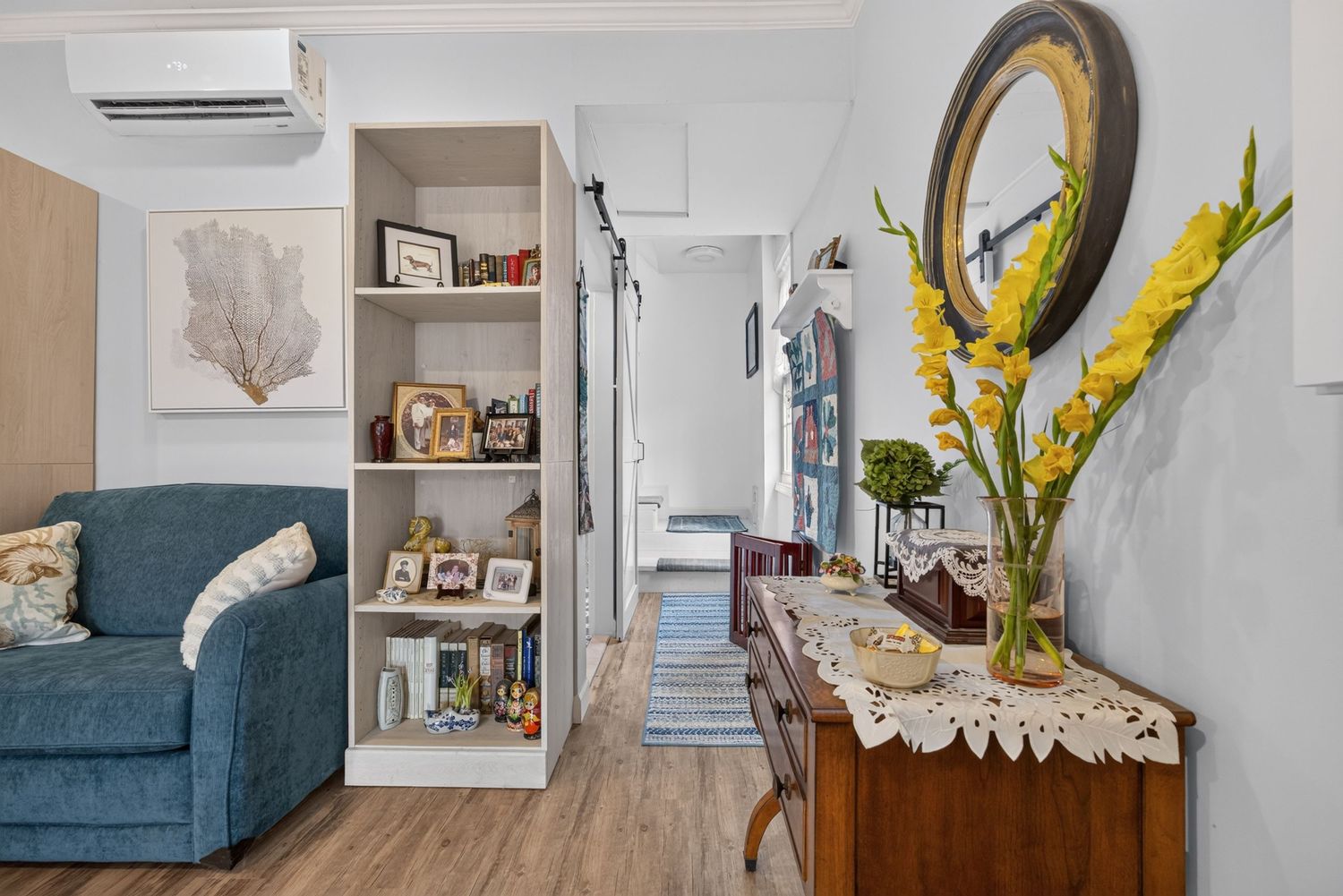 Cozy living room with blue sofa, white shelving unit, wooden sideboard with yellow flowers, and a round mirror on the wall.