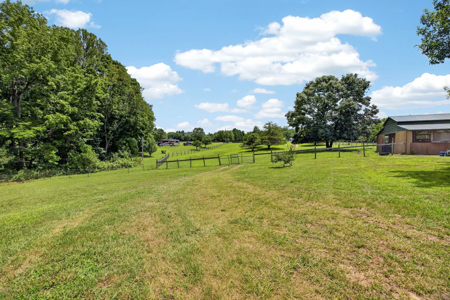 Lush green lawn with fenced pasture area, mature trees, and a partial view of a building on a sunny day.