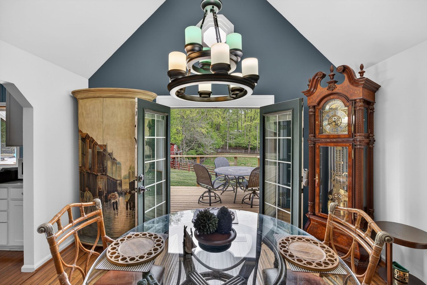 Elegant dining room with glass table, rattan chairs, grandfather clock, French doors to patio, and vaulted blue ceiling with chandelier.