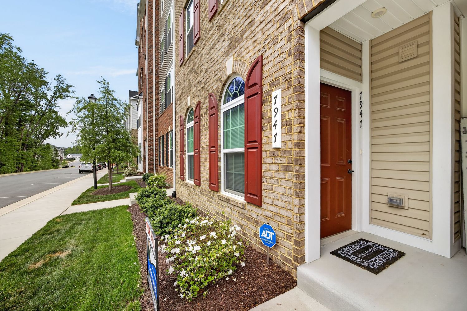 Brick townhouse exterior with red door, burgundy shutters, manicured landscaping, and a welcoming covered entryway.