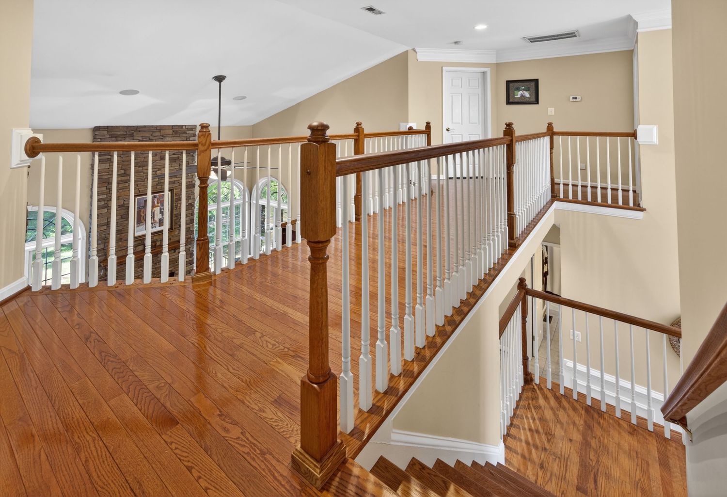 Elegant second-floor hallway with hardwood floors, wooden stair railings, white balusters, and open staircase in a luxury home.