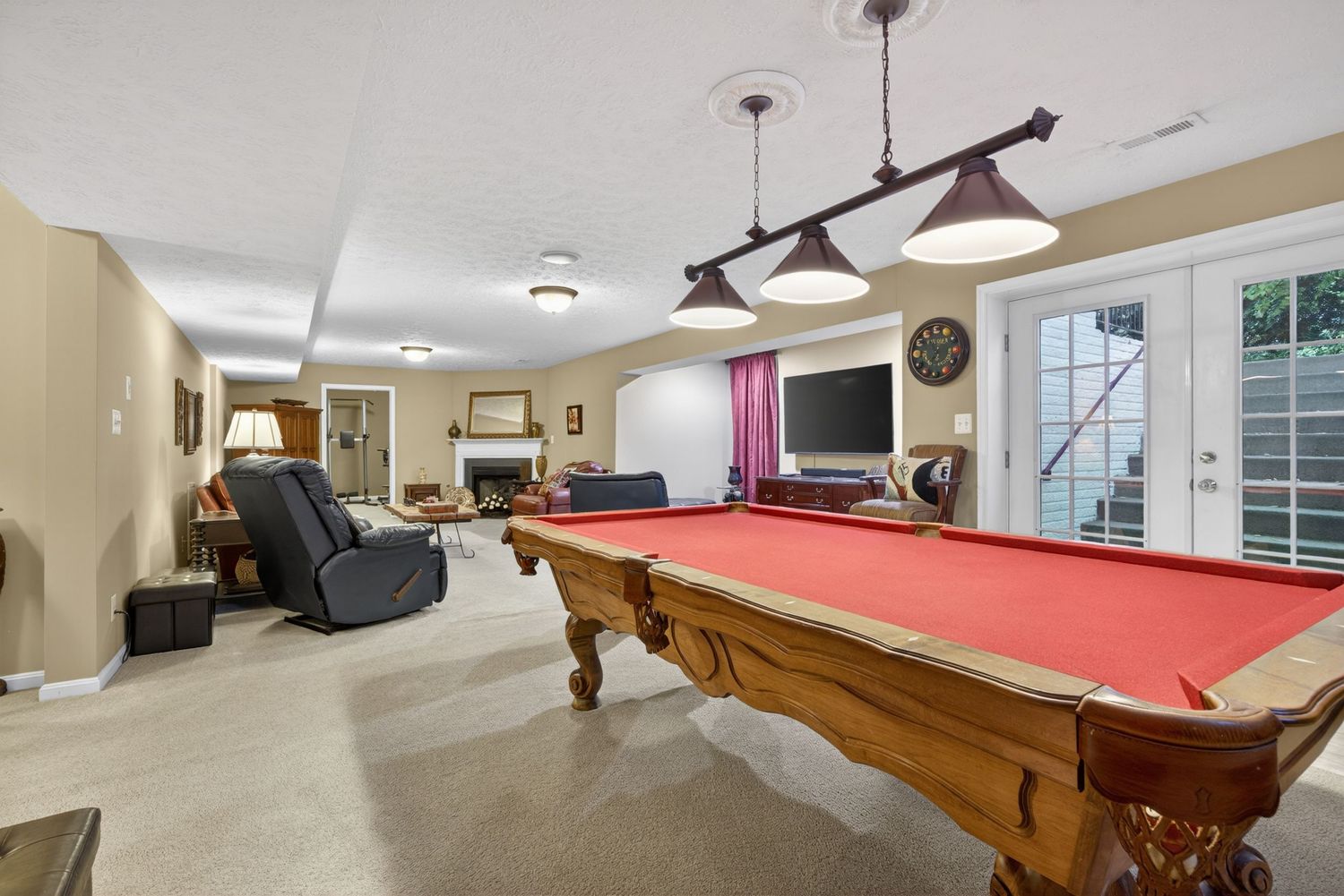 Spacious basement rec room featuring a wooden pool table with red felt, pendant lights, fireplace, and French doors.