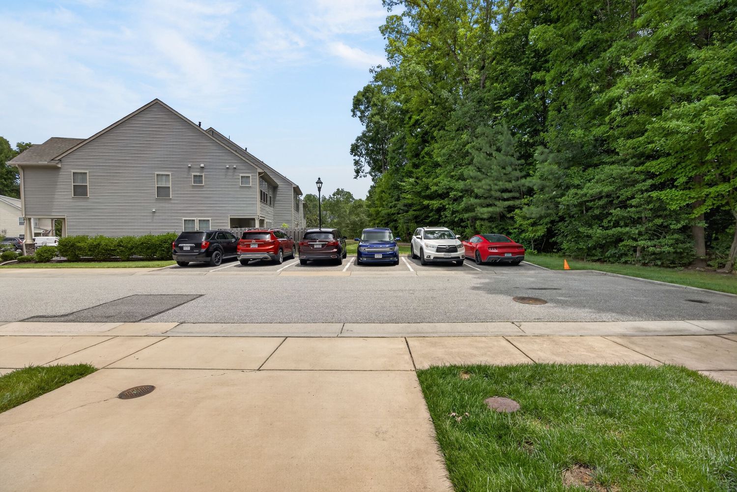 Suburban parking lot with multiple cars parked near a residential building surrounded by lush green trees on a sunny day.