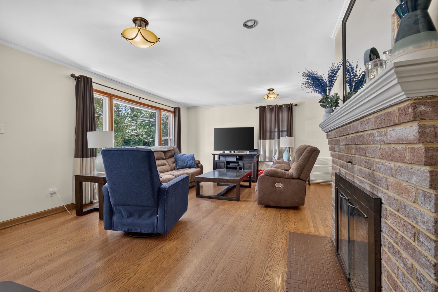 Bright living room with hardwood floors, brick fireplace, blue armchair, recliner, and large window with natural light.