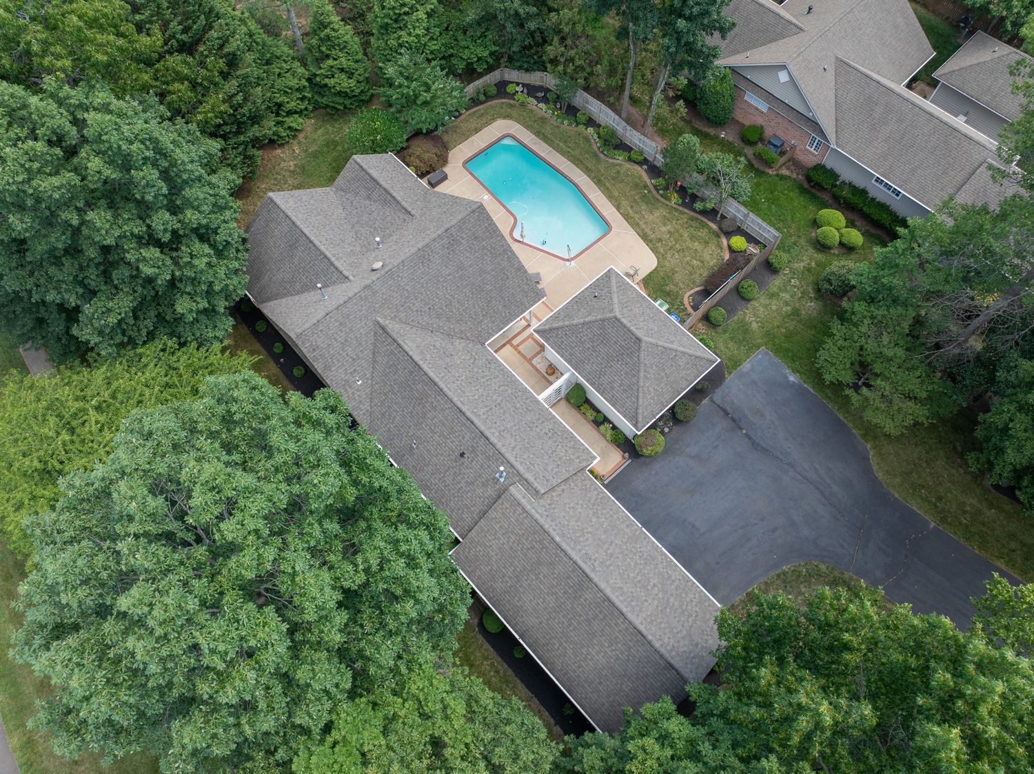 Aerial view of a large suburban home with a backyard swimming pool, surrounded by lush green trees and a paved driveway.