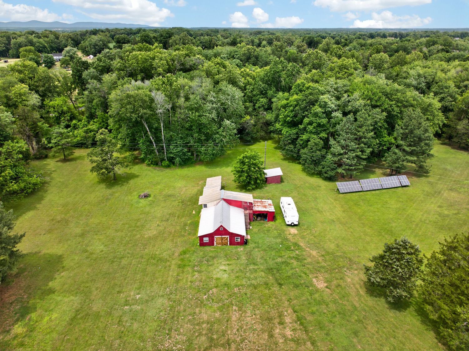 Aerial view of a rural property with red barn buildings surrounded by lush green trees and open fields with solar panels.
