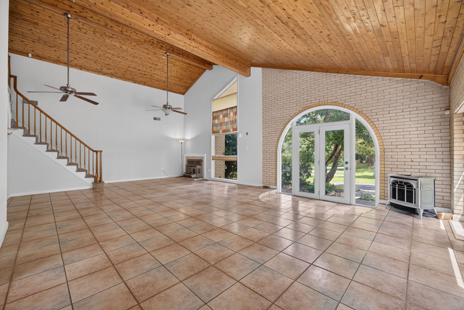 Spacious living room with vaulted wood ceiling, brick fireplace, tile floors, arched window, and French doors leading to backyard.