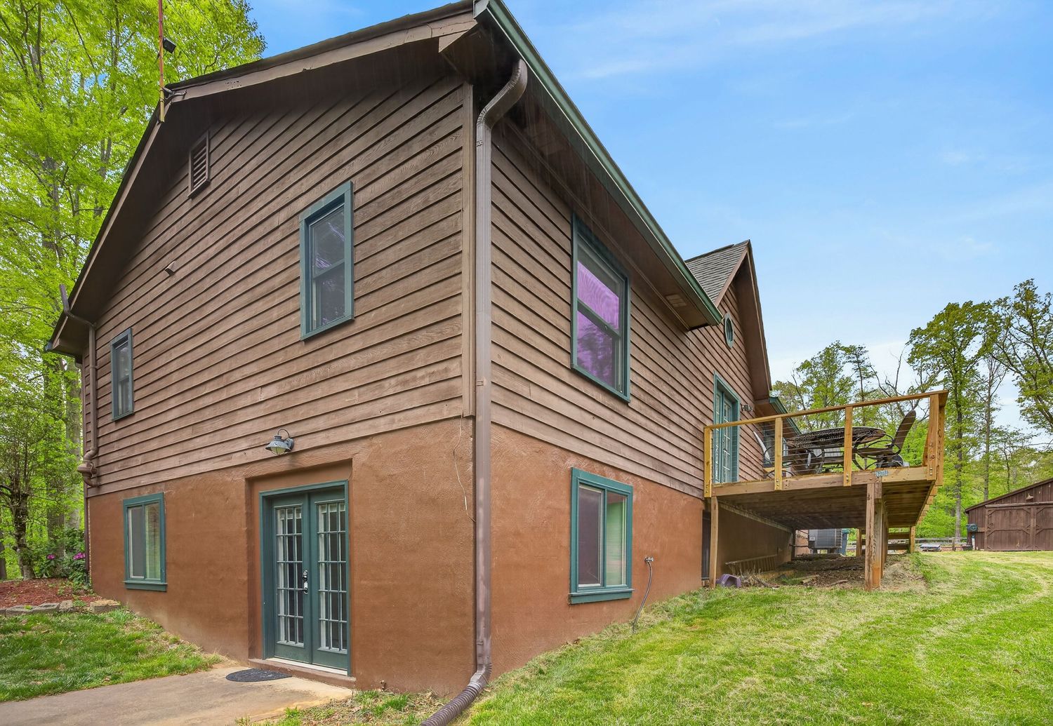 Brown two-story home with wood siding, green shutters, French doors, and a wooden deck surrounded by lush trees.