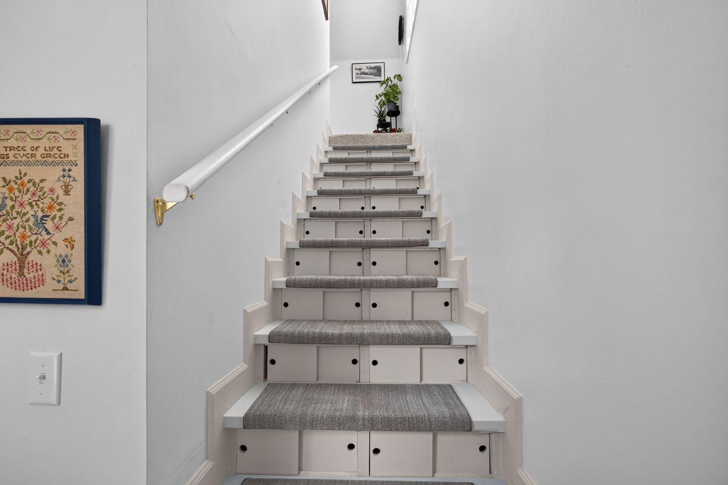 Modern staircase with gray carpet runners, white risers with built-in storage drawers, white walls, and decorative artwork.