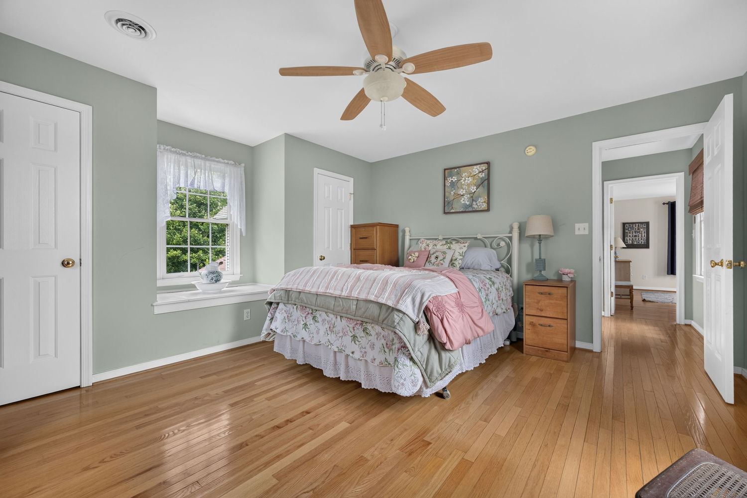Spacious bedroom with hardwood floors, sage green walls, ceiling fan, natural light from window, and cozy bedding.