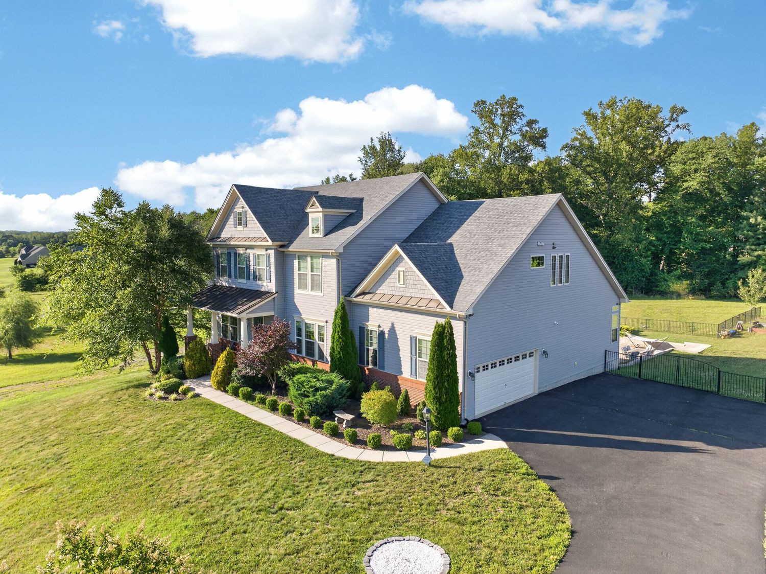 Aerial view of a large two-story colonial home with gray roof, attached garage, and lush green landscaping on a sunny day.