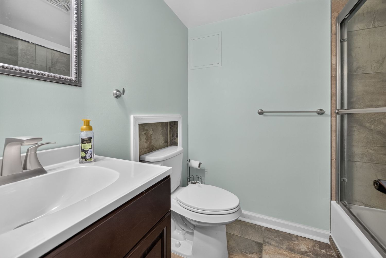 Modern bathroom with mint green walls, dark wood vanity, white toilet, and glass-enclosed bathtub with tile flooring.