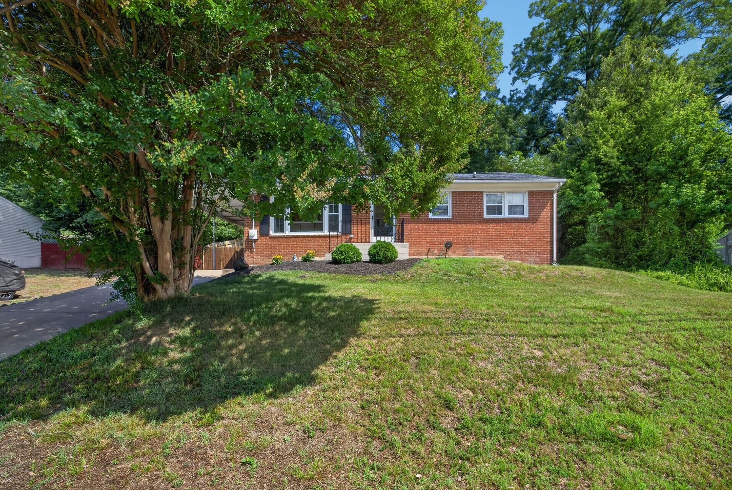Brick ranch home with large green lawn, mature trees, and landscaped shrubs on a sunny day.