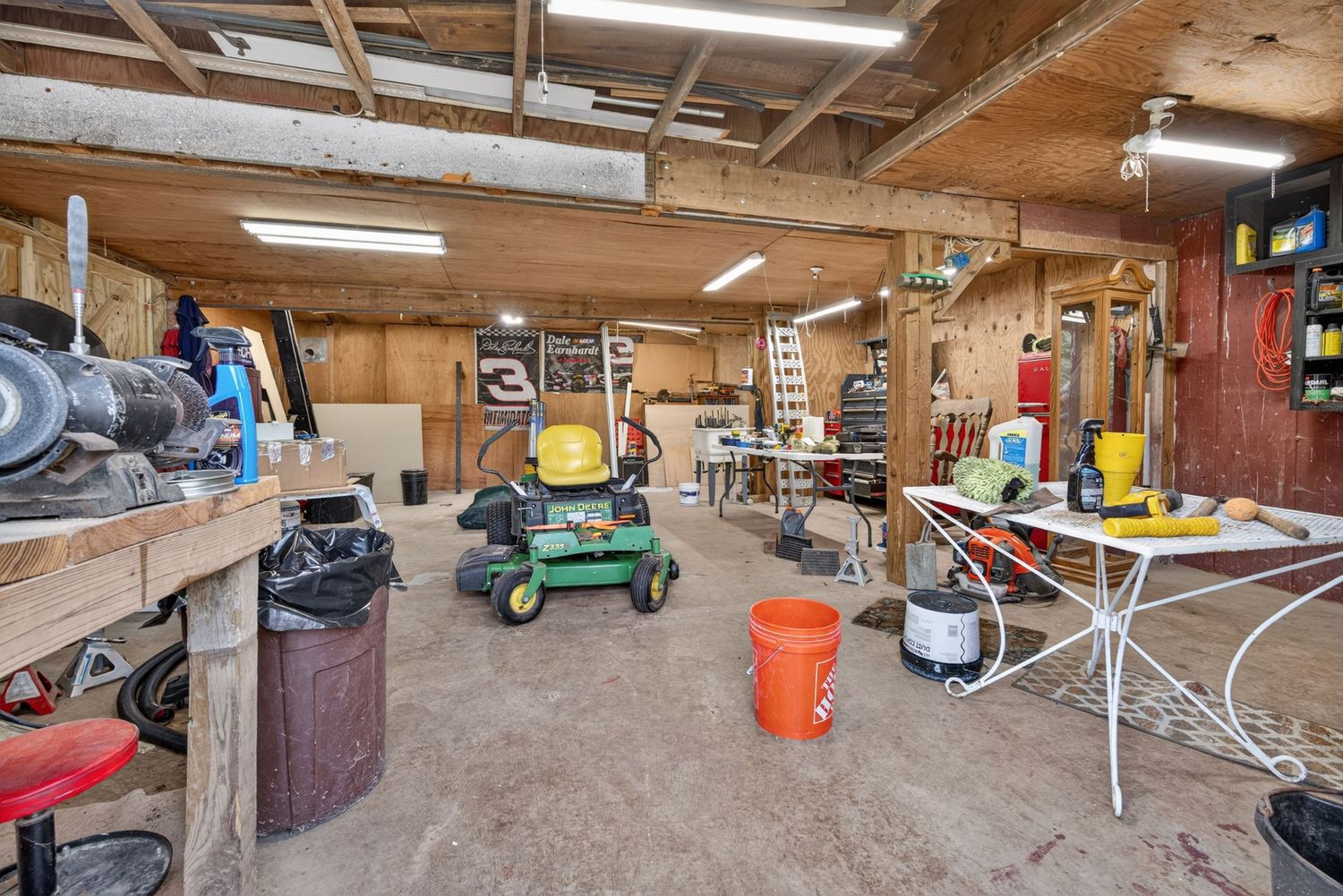 Spacious workshop garage with workbenches, lawn mower, tools, and storage shelving under fluorescent lighting.