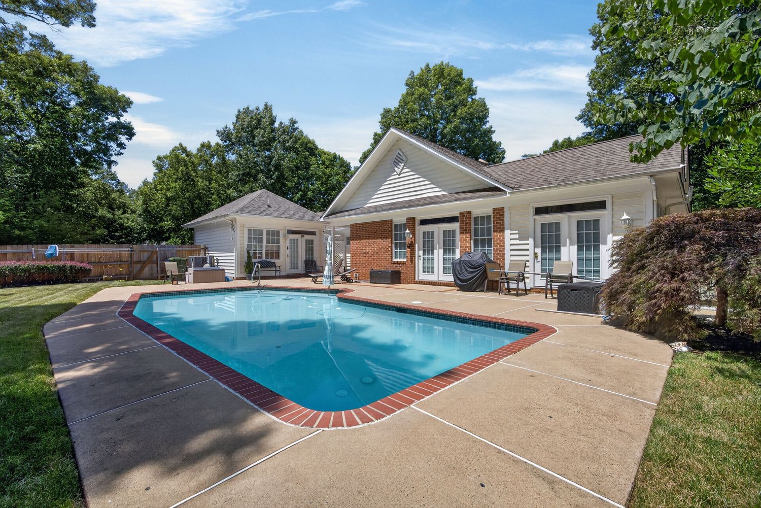 Backyard oasis featuring a kidney-shaped pool, brick patio, and white single-story home surrounded by mature trees.