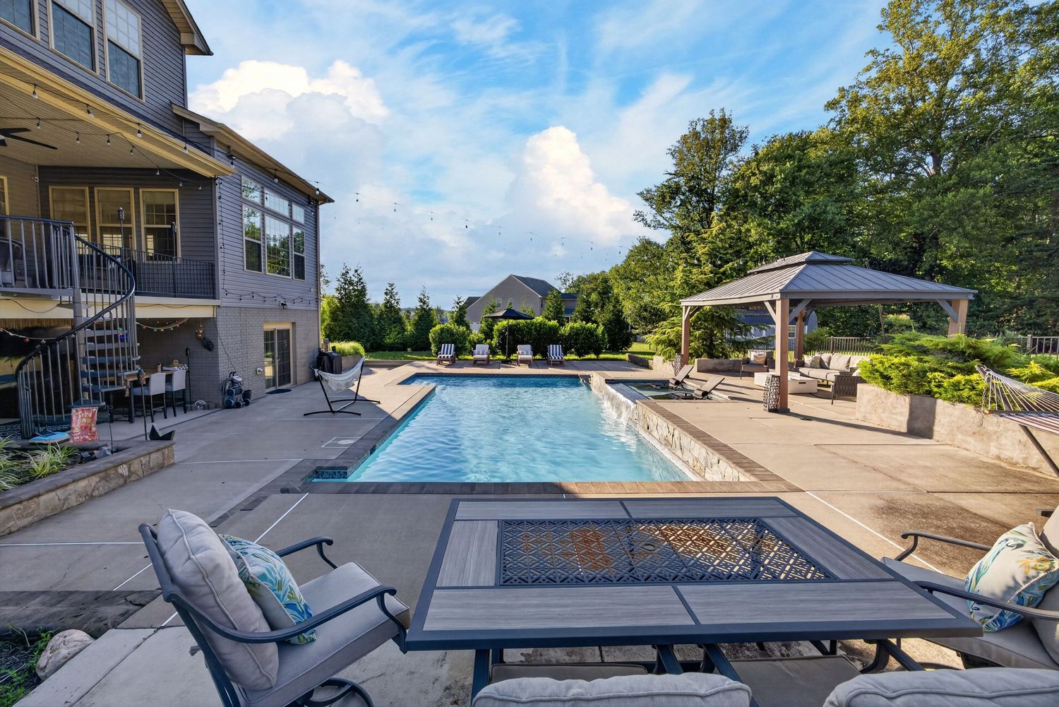 Luxury backyard patio with rectangular pool, gazebo, outdoor seating, fire pit table, and lush landscaping on a sunny day.