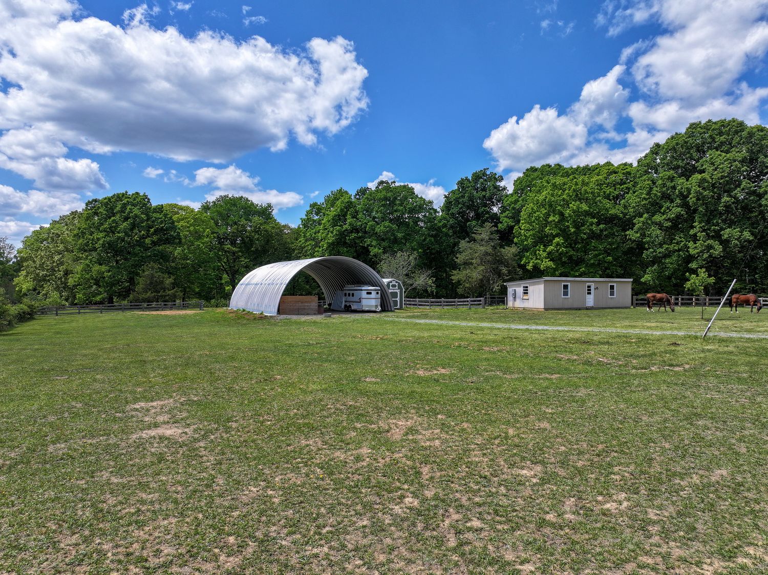 Rural property with quonset hut shelter, small outbuilding, and lush green trees under a blue sky with clouds.
