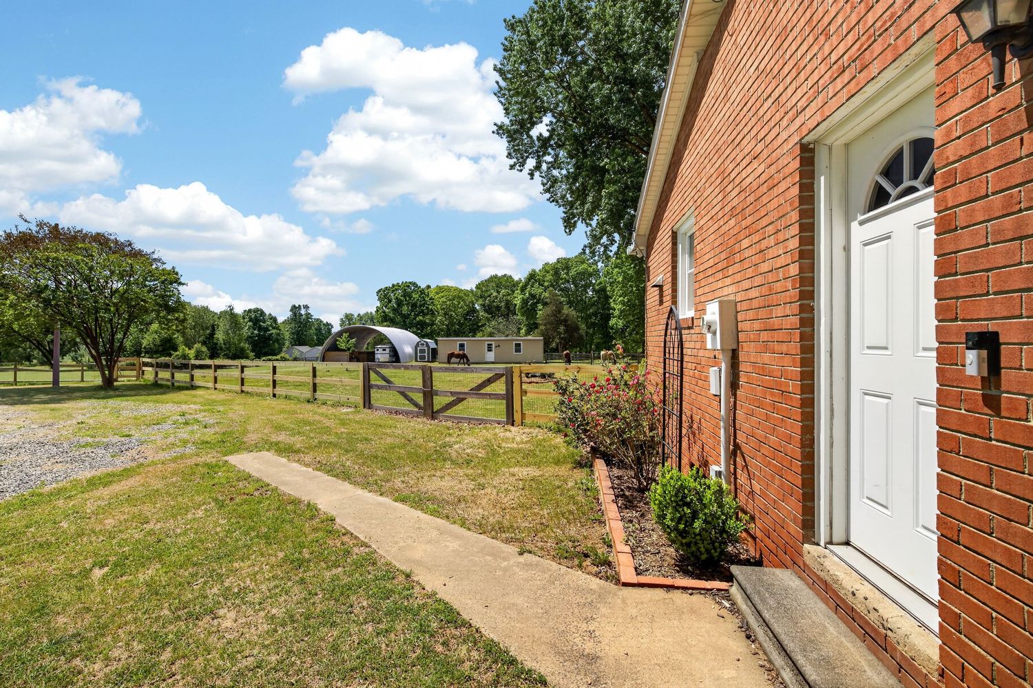 Brick home exterior with white front door, concrete walkway, and fenced pasture with trees under blue sky.