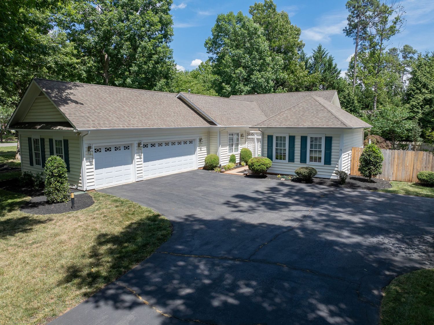 Ranch-style home with attached two-car garage, blue shutters, and lush green trees on a paved driveway.