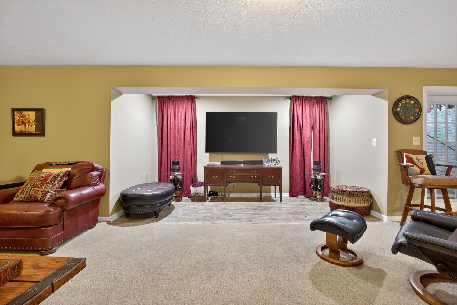Cozy living room with beige carpet, leather sofa, wall-mounted TV, red curtains, and warm neutral walls.