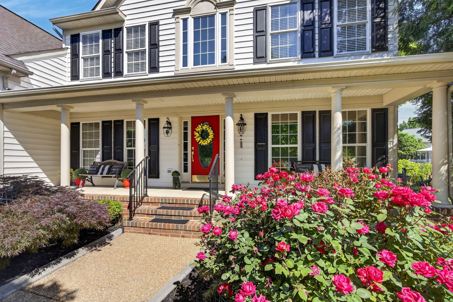 Charming colonial home with covered porch, black shutters, red wreath on door, and vibrant pink rose bushes in front yard.