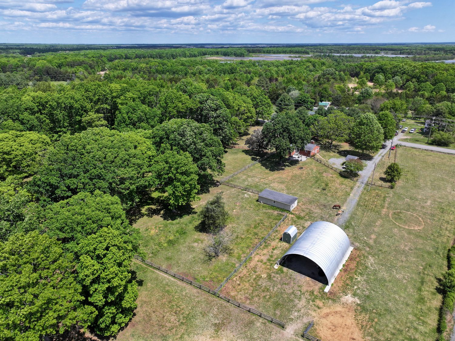 Aerial view of a rural property featuring a metal barn, open pastures, and lush green tree coverage under a partly cloudy sky.
