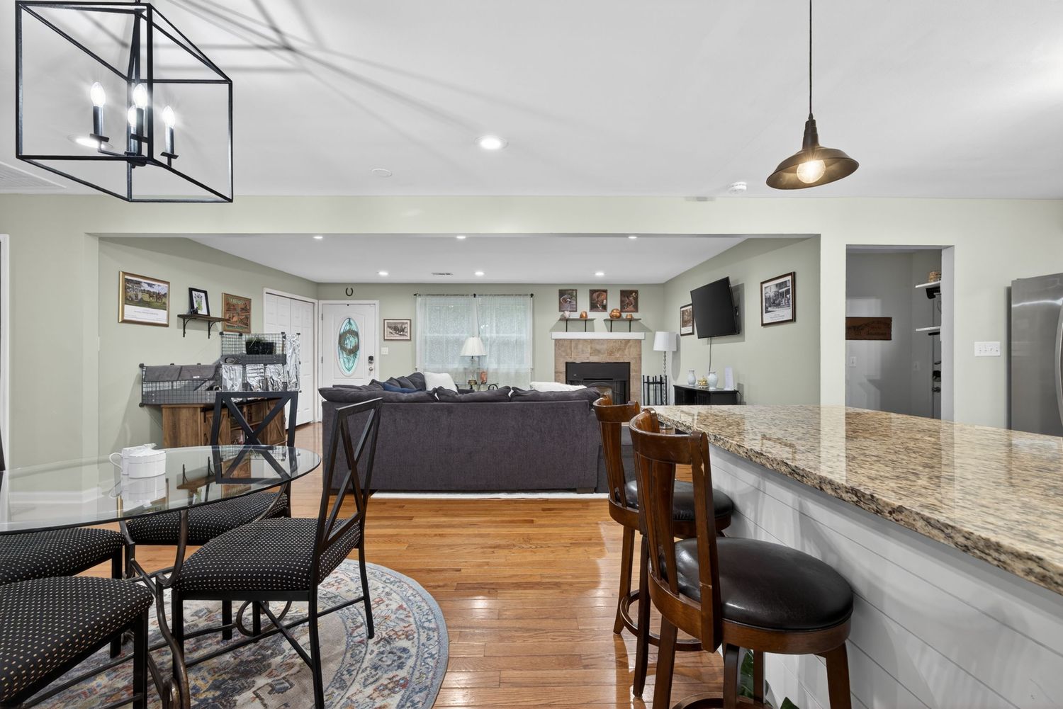 Open-concept kitchen and living area with granite countertop bar, hardwood floors, pendant lighting, and a cozy fireplace in background.