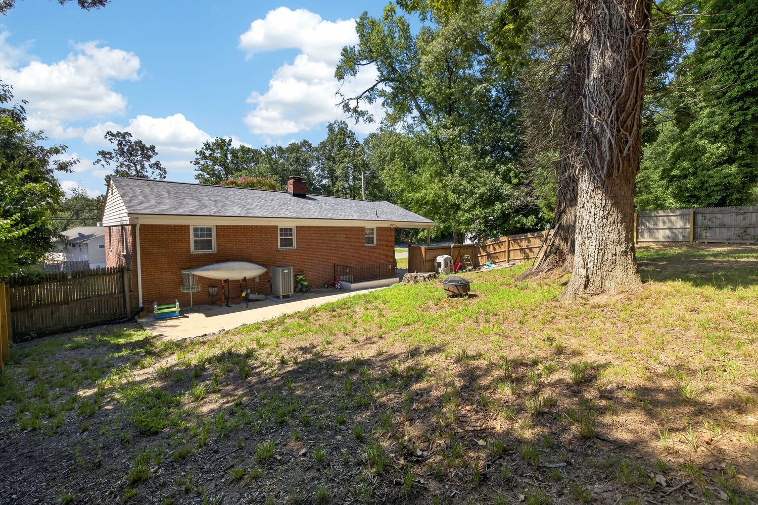 Brick ranch home with large backyard featuring a patio, mature trees, and wooden privacy fence on a sunny day.