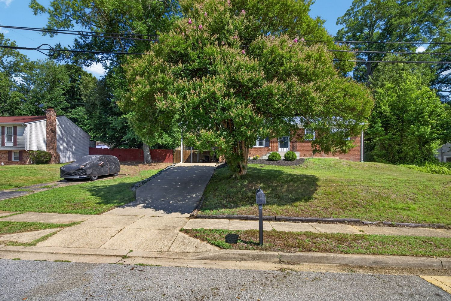 Suburban home with large tree in front yard, gravel driveway, and lush green lawn on a sunny day.