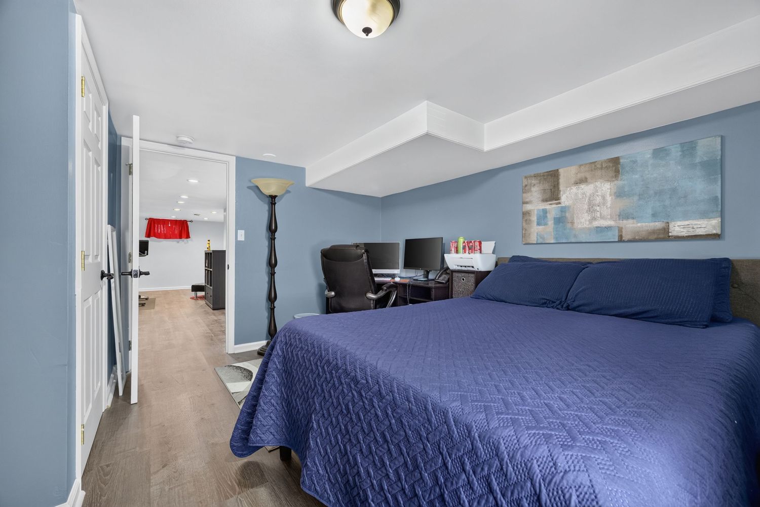 Spacious bedroom with blue bedding, tray ceiling, hardwood floors, and a home office area visible in the corner.