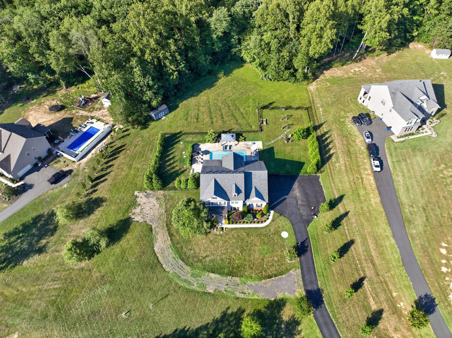 Aerial view of a suburban home with a pool, circular driveway, and lush green lawn surrounded by dense trees.