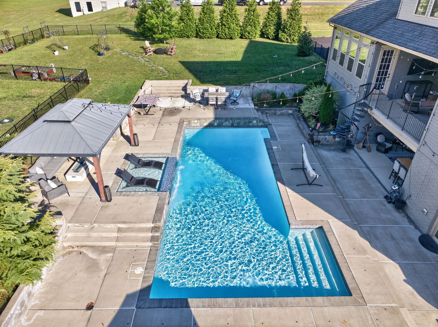 Aerial view of a rectangular backyard swimming pool with steps, patio seating, gazebo, and lush green lawn.