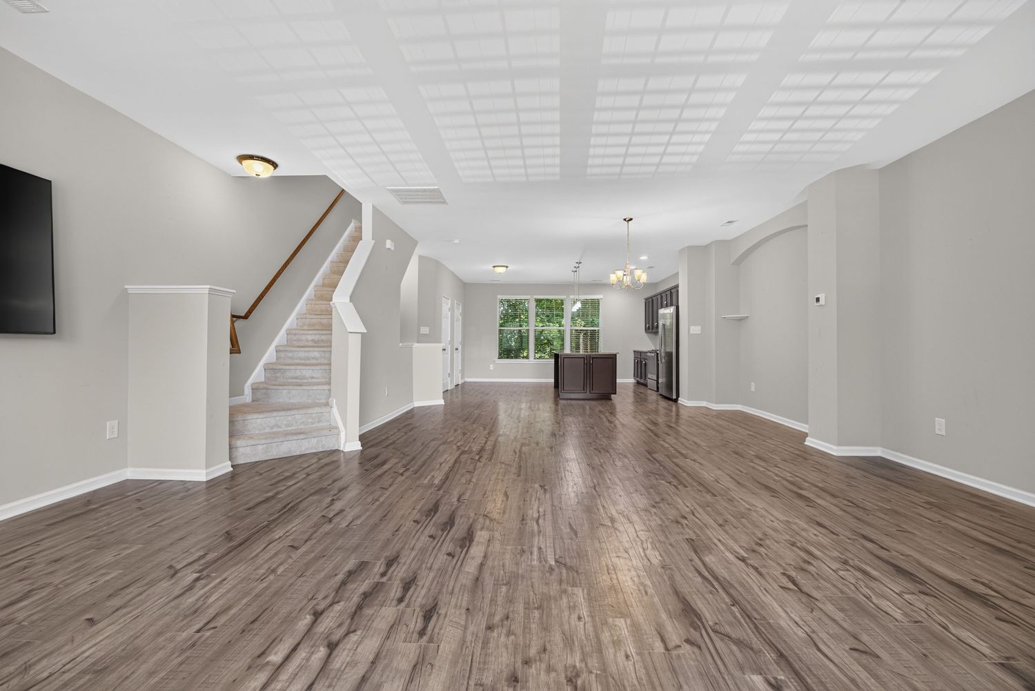Spacious open-concept living area with dark hardwood floors, white trim, coffered ceiling, staircase, and chandelier in background.