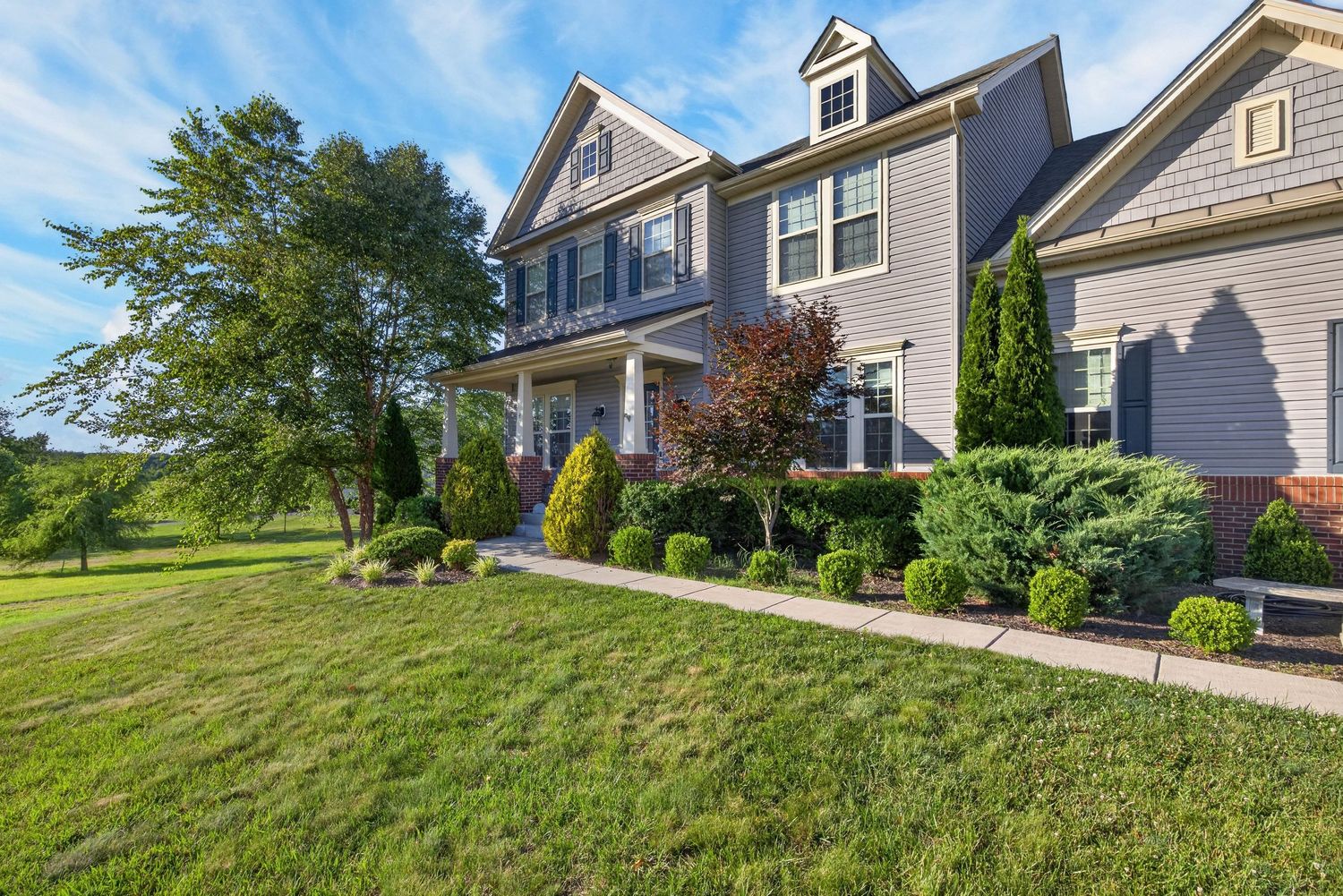 A large two-story gray colonial home with a covered porch, lush green lawn, and manicured landscaping on a sunny day.