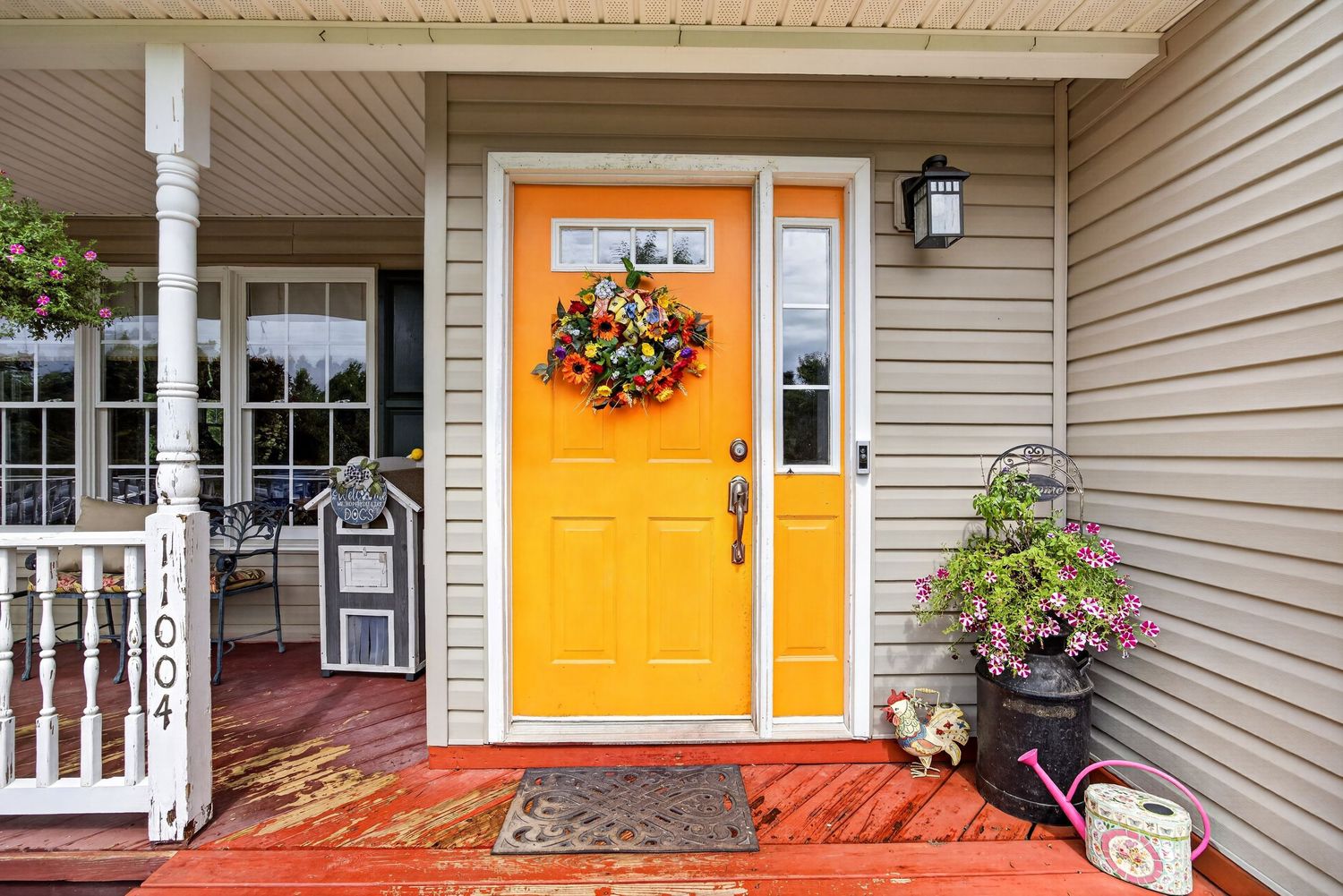 Bright yellow front door with colorful wreath on gray house with white porch railing and potted plants.