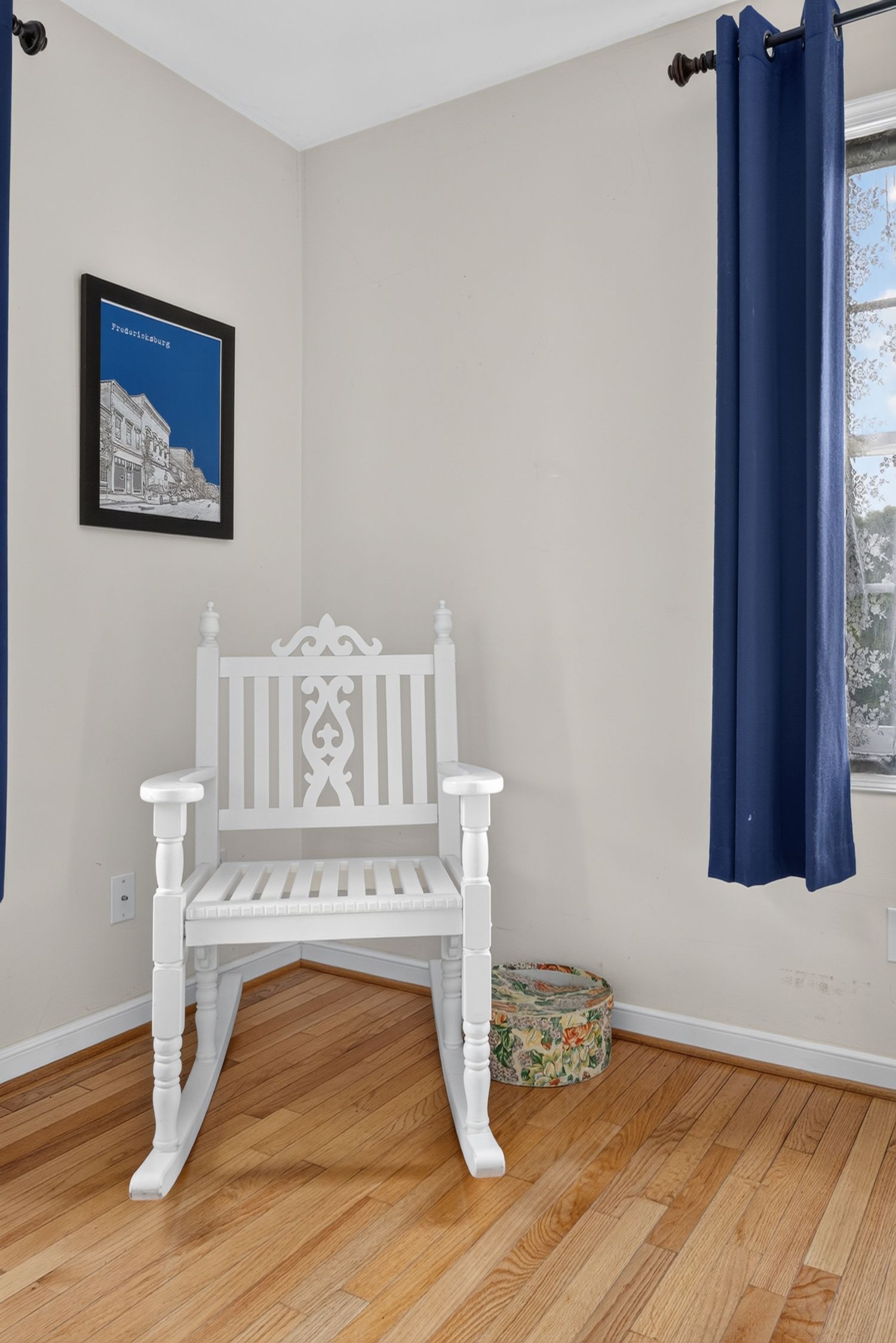 White ornate rocking chair on hardwood floors in a cozy corner with blue curtains and a framed city art print.