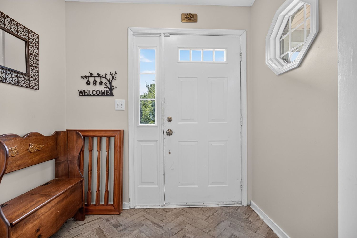 Bright entryway with white front door, sidelights, herringbone tile floors, wooden bench, and decorative welcome sign.