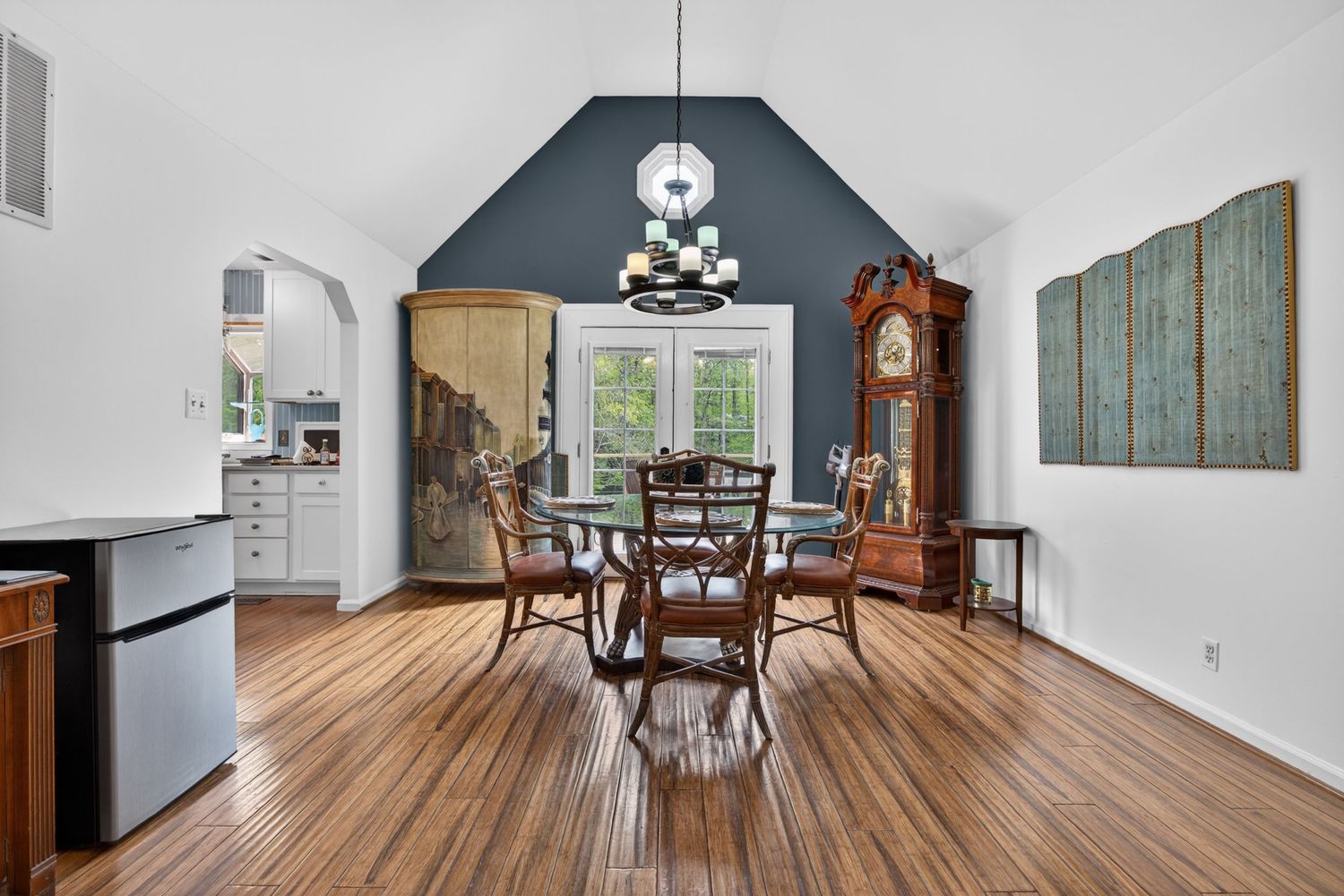 Dining room with hardwood floors, round glass table, vaulted ceiling with chandelier, blue accent wall, and arched doorway.