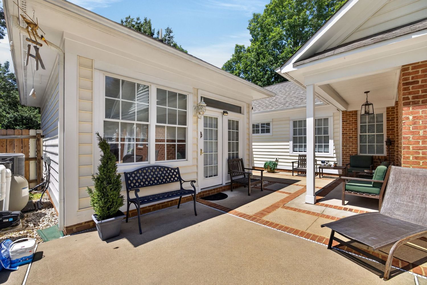Sunny patio with black bench, outdoor seating, French doors, white siding, and brick accents surrounded by lush greenery.