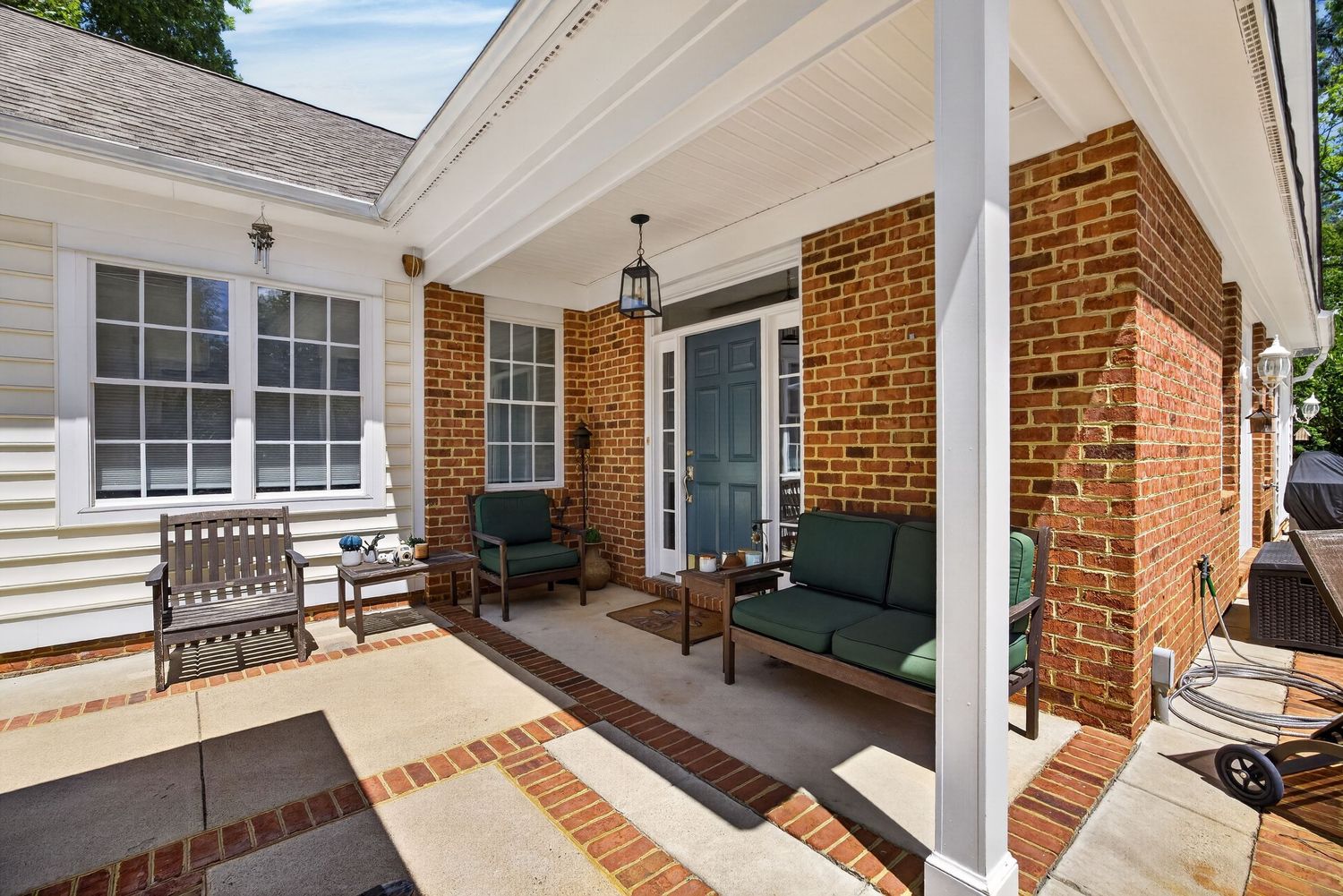 Charming brick home covered porch with green outdoor furniture, lantern light fixture, and white columns on a patio.