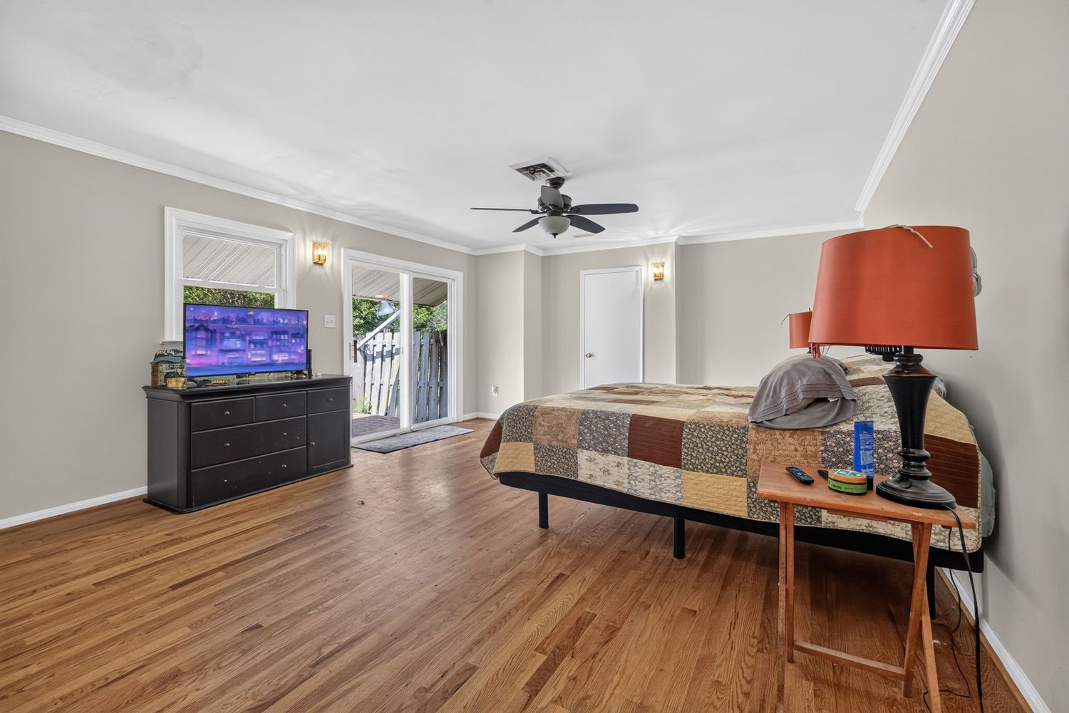 Spacious bedroom with hardwood floors, crown molding, ceiling fan, French doors, a patchwork bed, dresser with TV, and side table.
