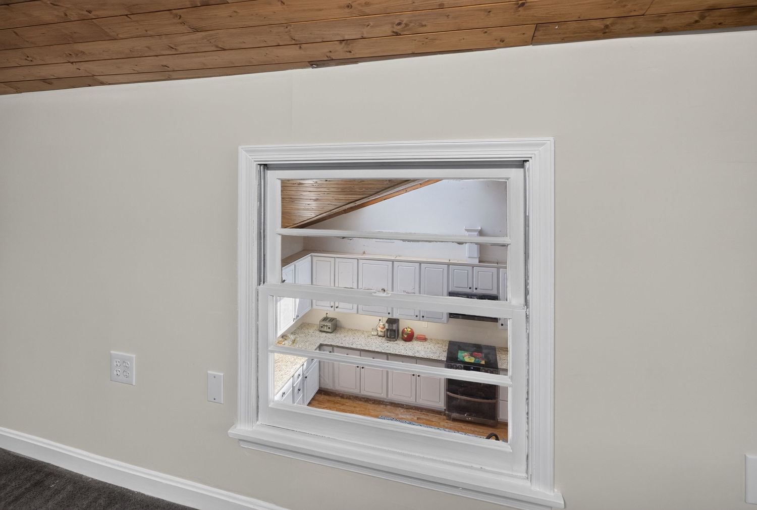 Interior pass-through window revealing a white kitchen with cabinetry and wood ceiling beyond a neutral wall.