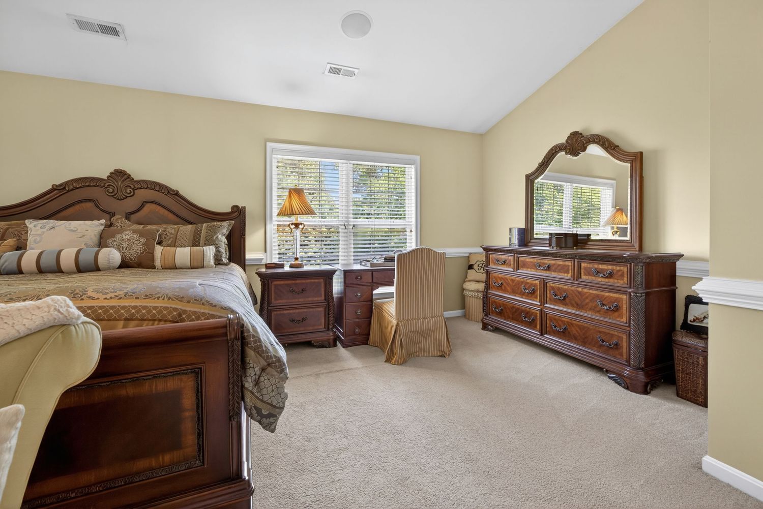 Spacious bedroom with dark wood furniture set, beige carpet, light yellow walls, and natural light from two windows.