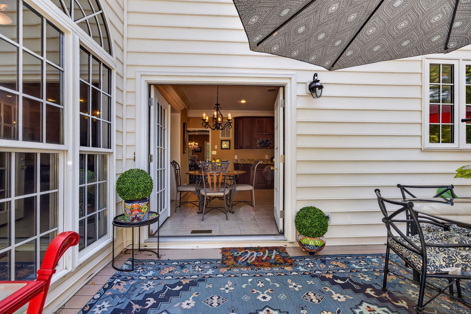 Charming home patio entrance with blue floral rug, topiary plants, and open door revealing elegant dining area inside.