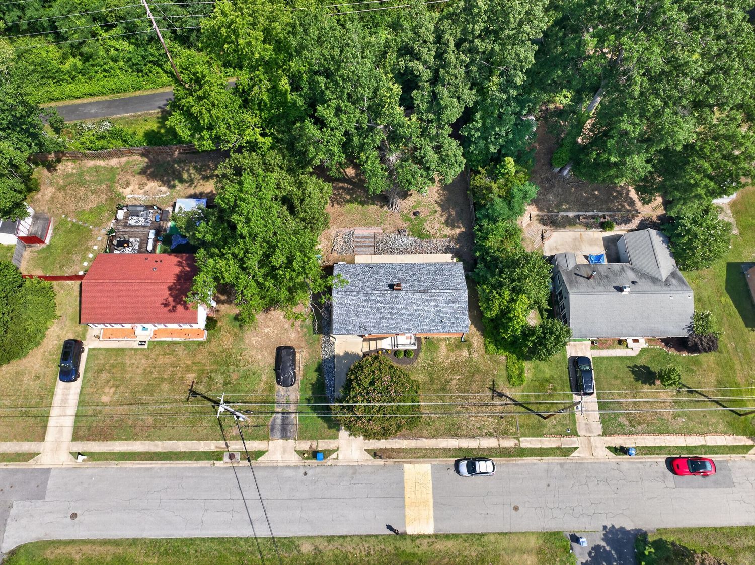 Aerial drone view of suburban residential properties with tree-lined street, multiple homes, and green lawns on a sunny day.