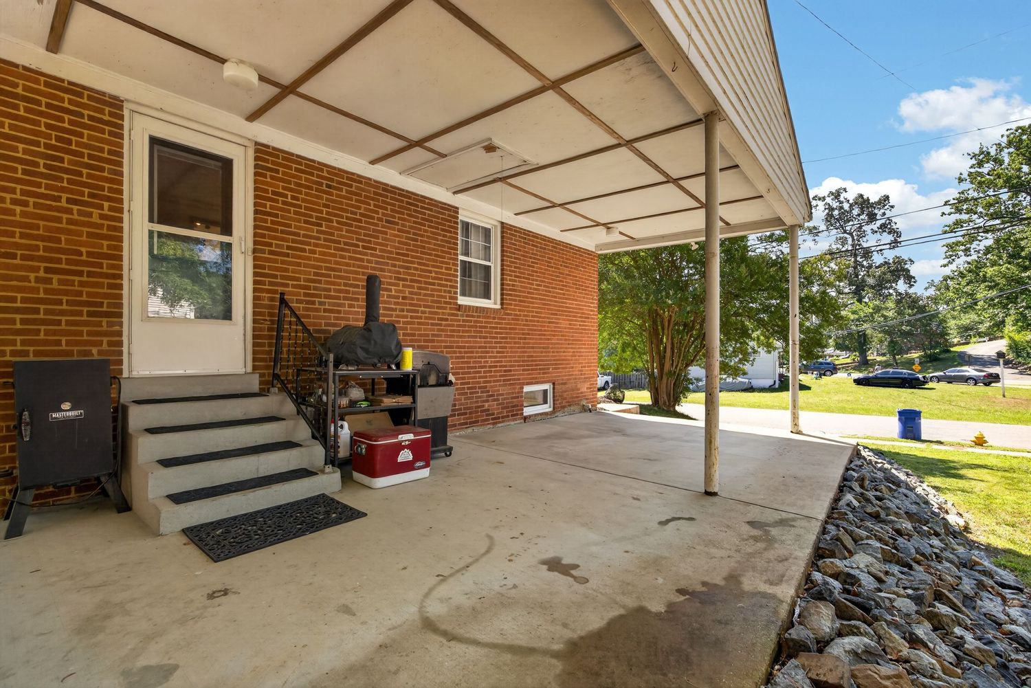 Covered concrete patio of a brick home with outdoor grill, steps leading to back door, and lush green trees in background.