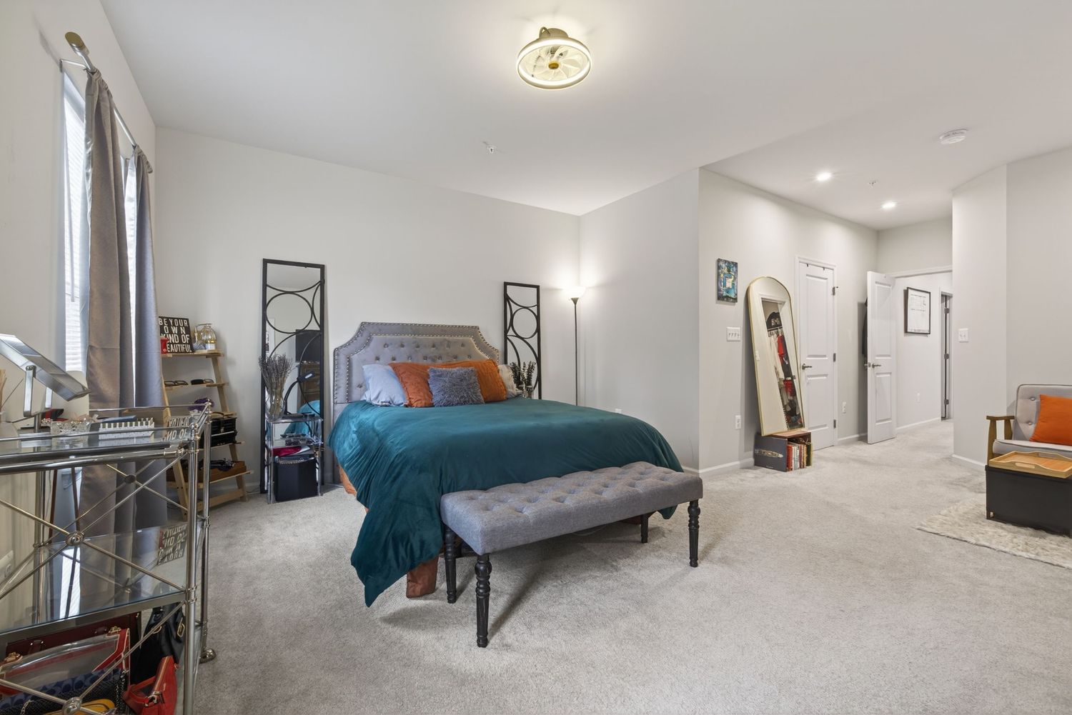 Spacious bedroom with teal bedspread, gray tufted bench, plush carpet, and bright natural light from large windows.