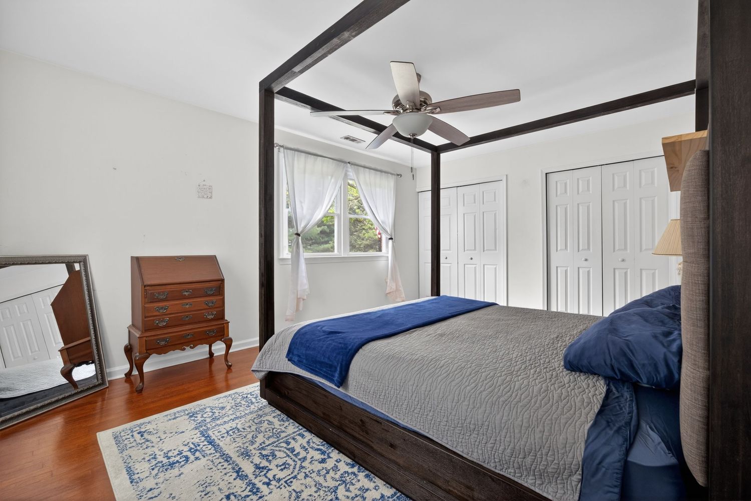 Elegant four-poster bed bedroom with hardwood floors, ceiling fan, blue bedding, antique dresser, and white closet doors.
