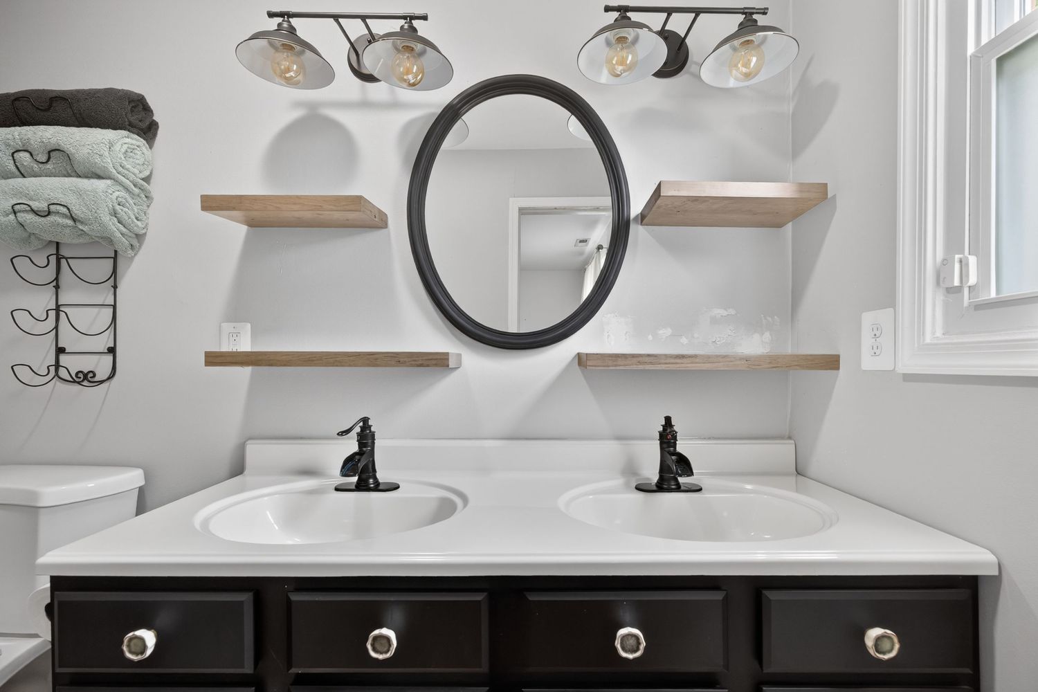 Modern double vanity bathroom with dark cabinets, oval mirror, floating wood shelves, and dual undermount sinks with black faucets.
